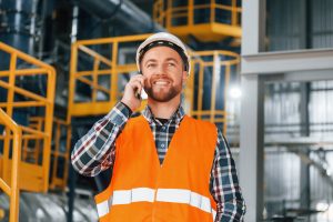 Talking by phone. Construction worker in uniform is in the factory.