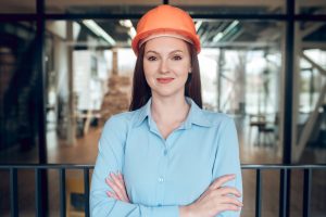 All perfectly. Optimistic young smiling woman in safety helmet standing with crossed arms during working hours indoors