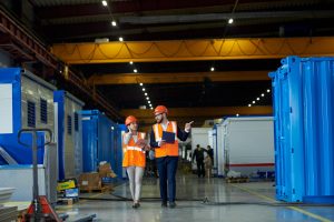 Full length  portrait of bearded businessman wearing hardhat talking to female factory worker while discussing production in workshop, copy space