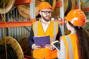 Portrait of modern bearded businessman wearing hardhat listening to factory employee while standing in workshop and holding clipboard, copy space